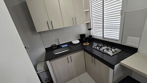 A compact kitchen with light wood cabinets, a black countertop, a gas stove, and a window with blinds. - Apartaestudios San Fernando (Cali)