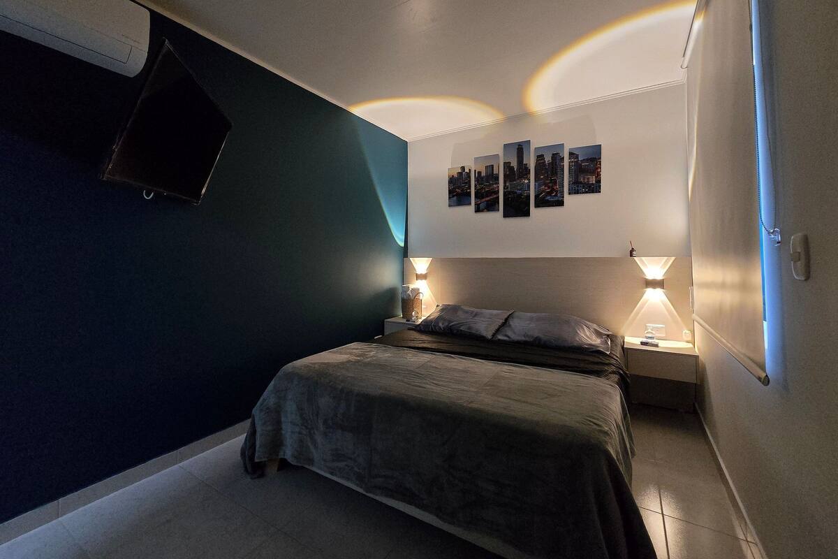A bedroom with a bed, two bedside tables, a wall-mounted TV, and a cityscape artwork.