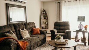 A living room with a dark brown sofa, a matching armchair, a wooden coffee table, and a decorative shelf with plants and framed pictures. - The Urban Oasis (Tignish)