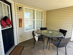 A covered patio with a round glass table and four chairs, a black door, and a red decorative item on the door.