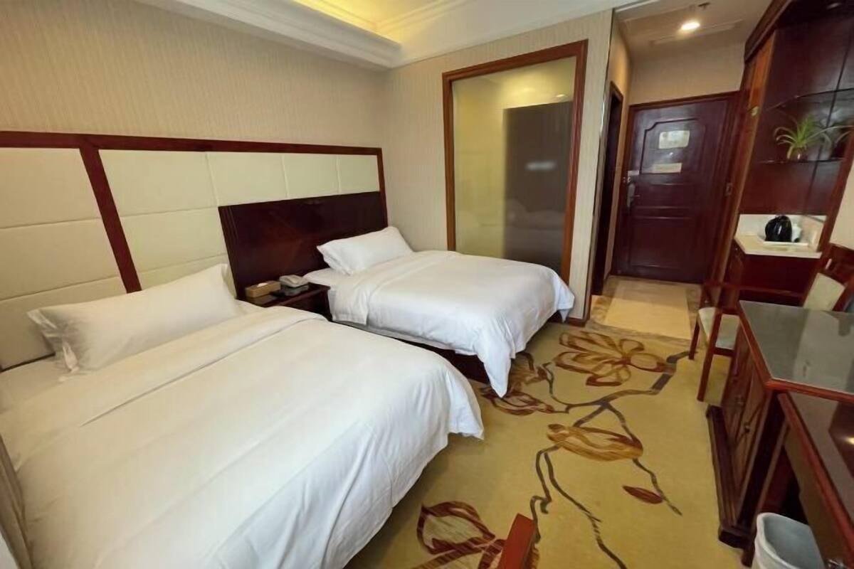 A hotel room with two beds, a wooden headboard, a nightstand, a desk, a chair, a mirror, a wardrobe, and a door.