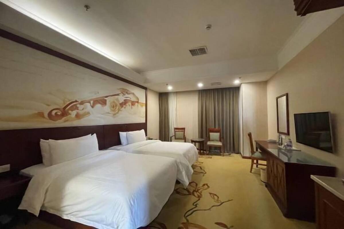 A hotel room with two beds, a desk, a chair, a television, and a large painting on the wall.