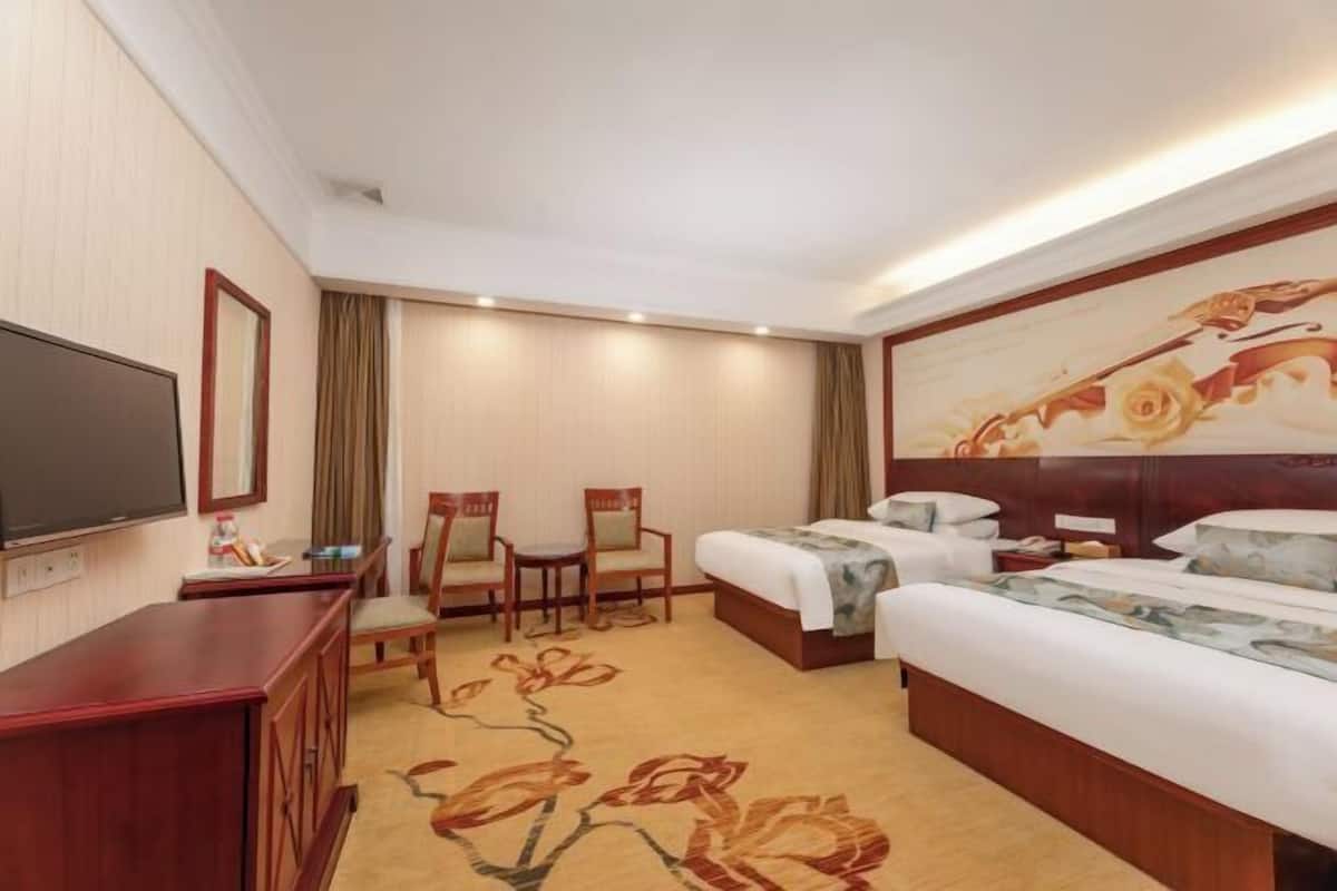 A hotel room with a large bed, a smaller bed, a desk with a flat-screen TV, two chairs, and a decorative wall painting.