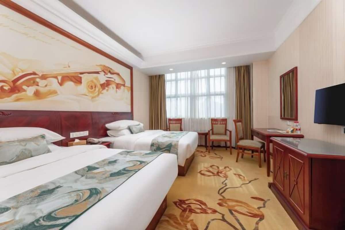 A hotel room with two beds, a desk, a chair, a TV, and a large floral painting on the wall.
