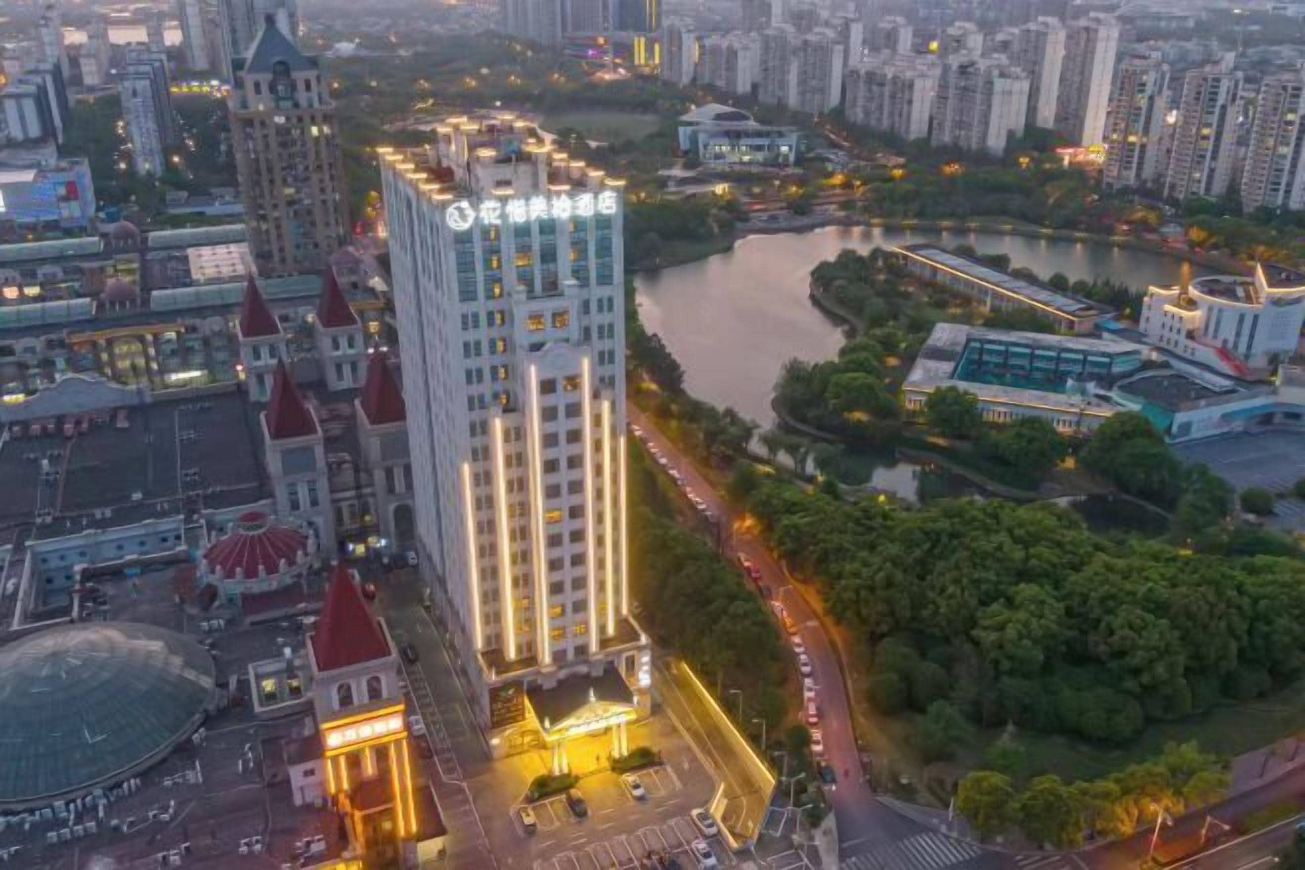 Un hotel moderno de gran altura con una entrada destacada, rodeado por un río y vegetación, junto a un edificio histórico con tejado rojo.