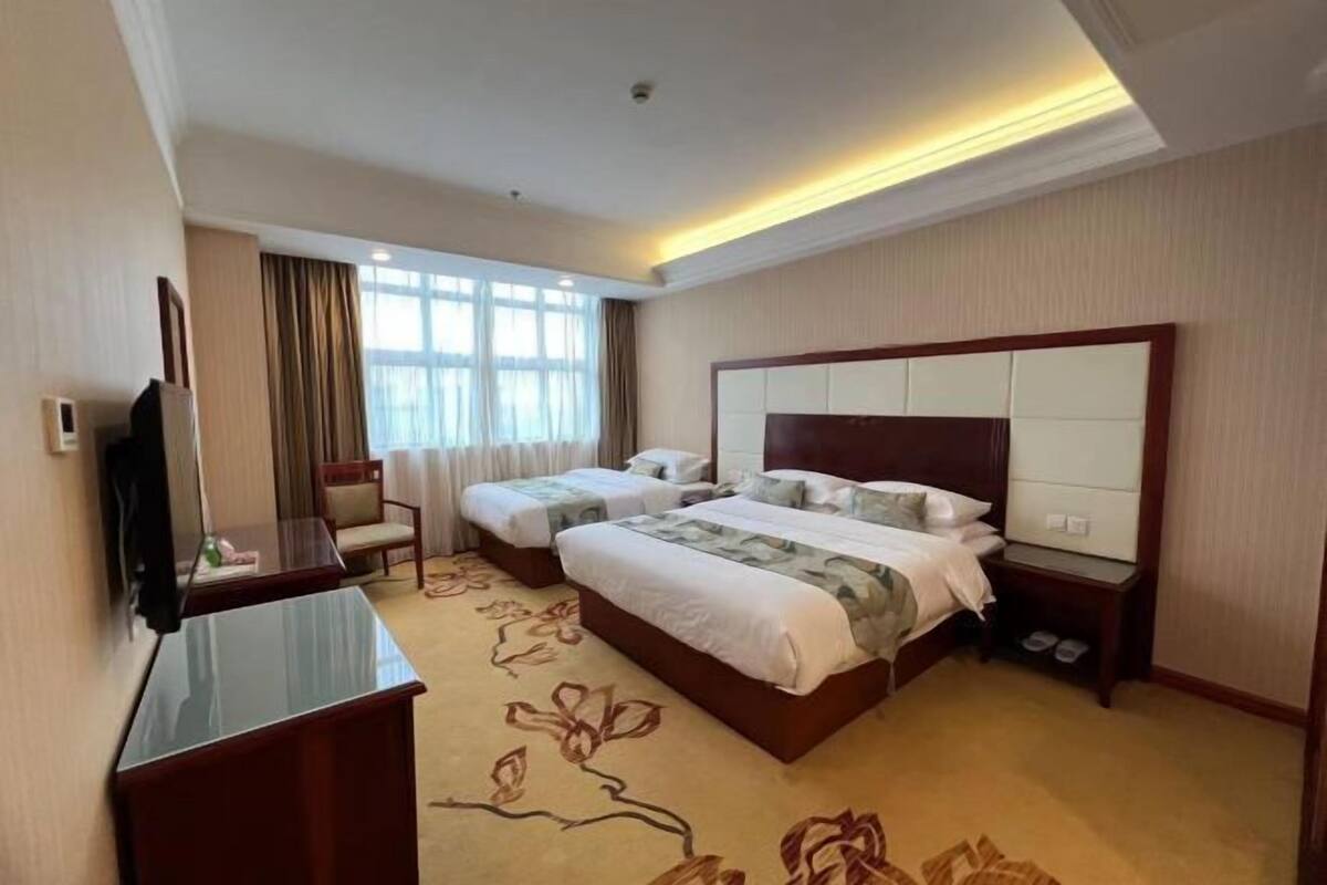 A hotel room with two beds, a desk, a TV, and a large window with curtains.
