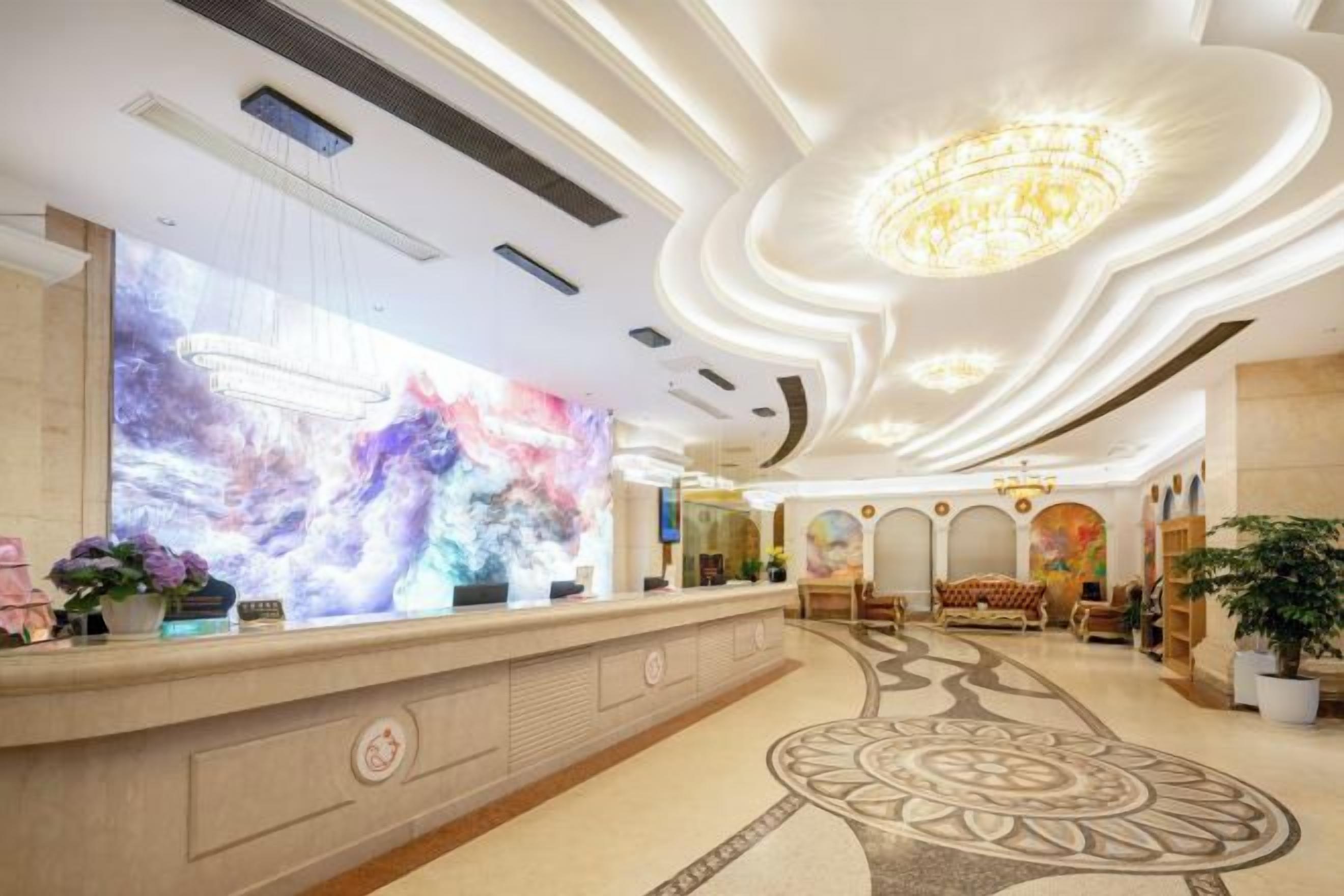 A hotel lobby with a reception desk, a large abstract mural, a chandelier, and a patterned carpet.