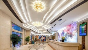 A hotel lobby with a central reception desk, a patterned floor, and a large chandelier.