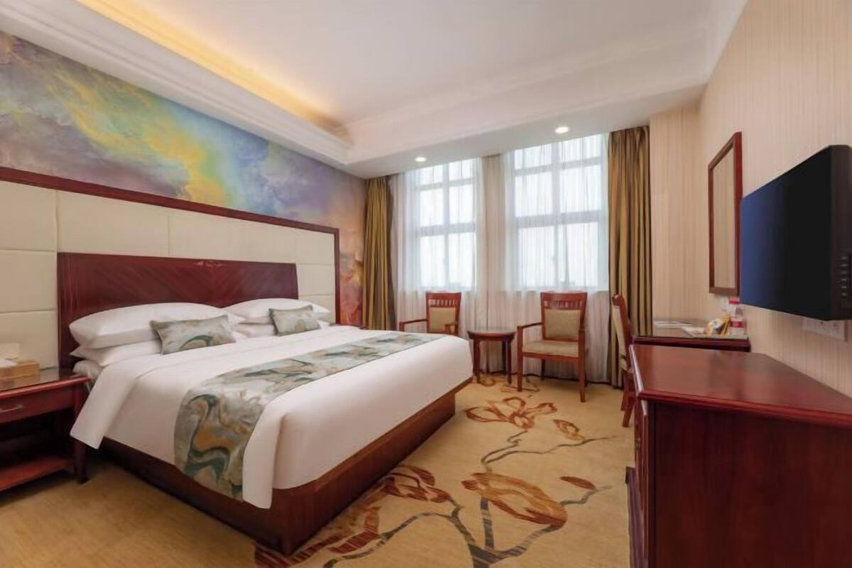A hotel room with a large bed, a desk, a chair, a TV, and a painting on the wall.