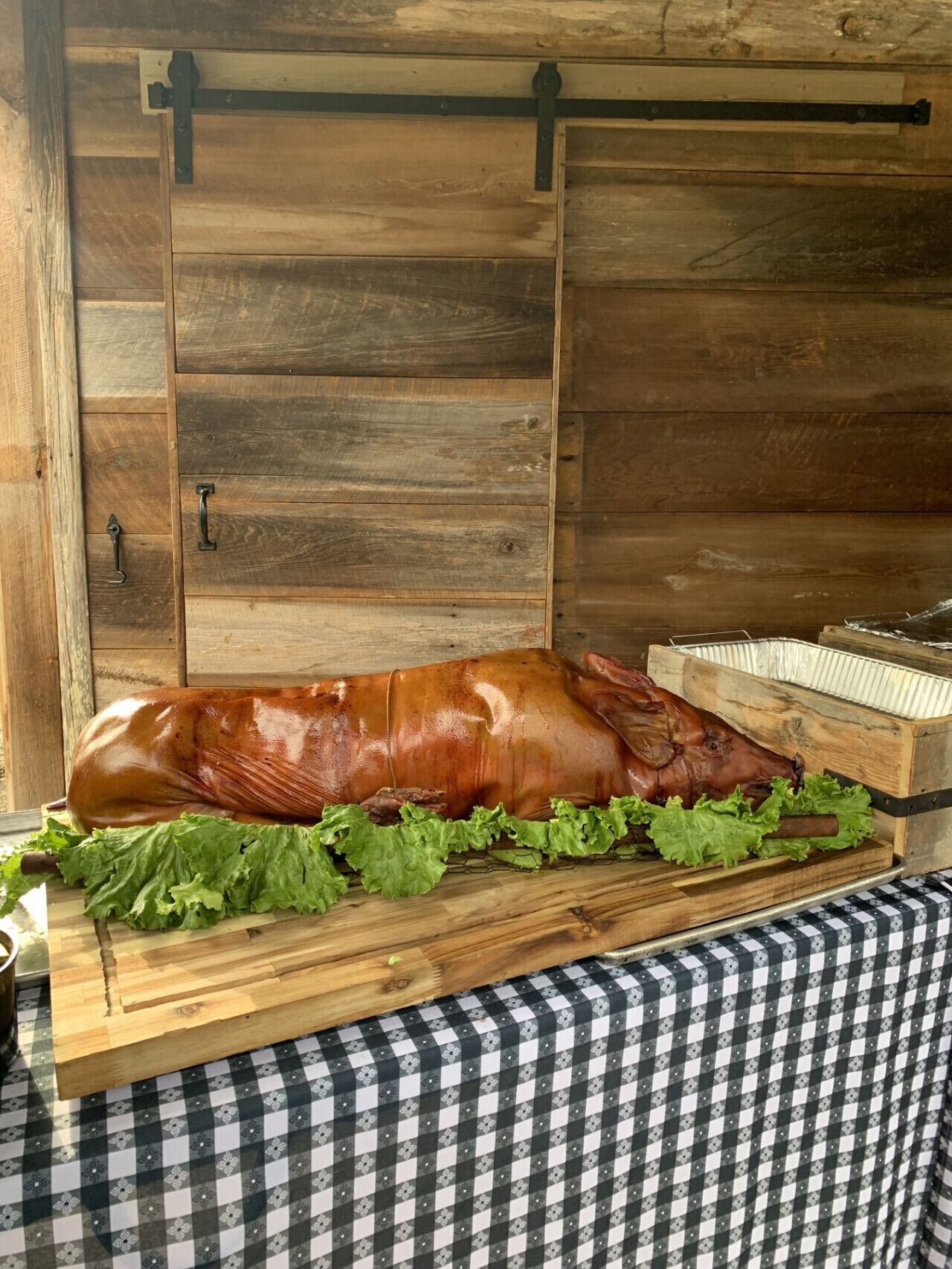 Un cerdo asado sobre una mesa de madera con mantel a cuadros, frente a una pared de madera rústica con una puerta corrediza.