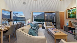 A modern living room with a sofa, dining table, and chairs, large windows with scenic views, and decorative lighting. - Swallowdale | A home away from home, close to town (Queenstown)
