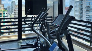 Un gimnasio con cintas de correr y equipamiento de ejercicio, ventanas amplias con vista a la ciudad y un ventilador de techo.