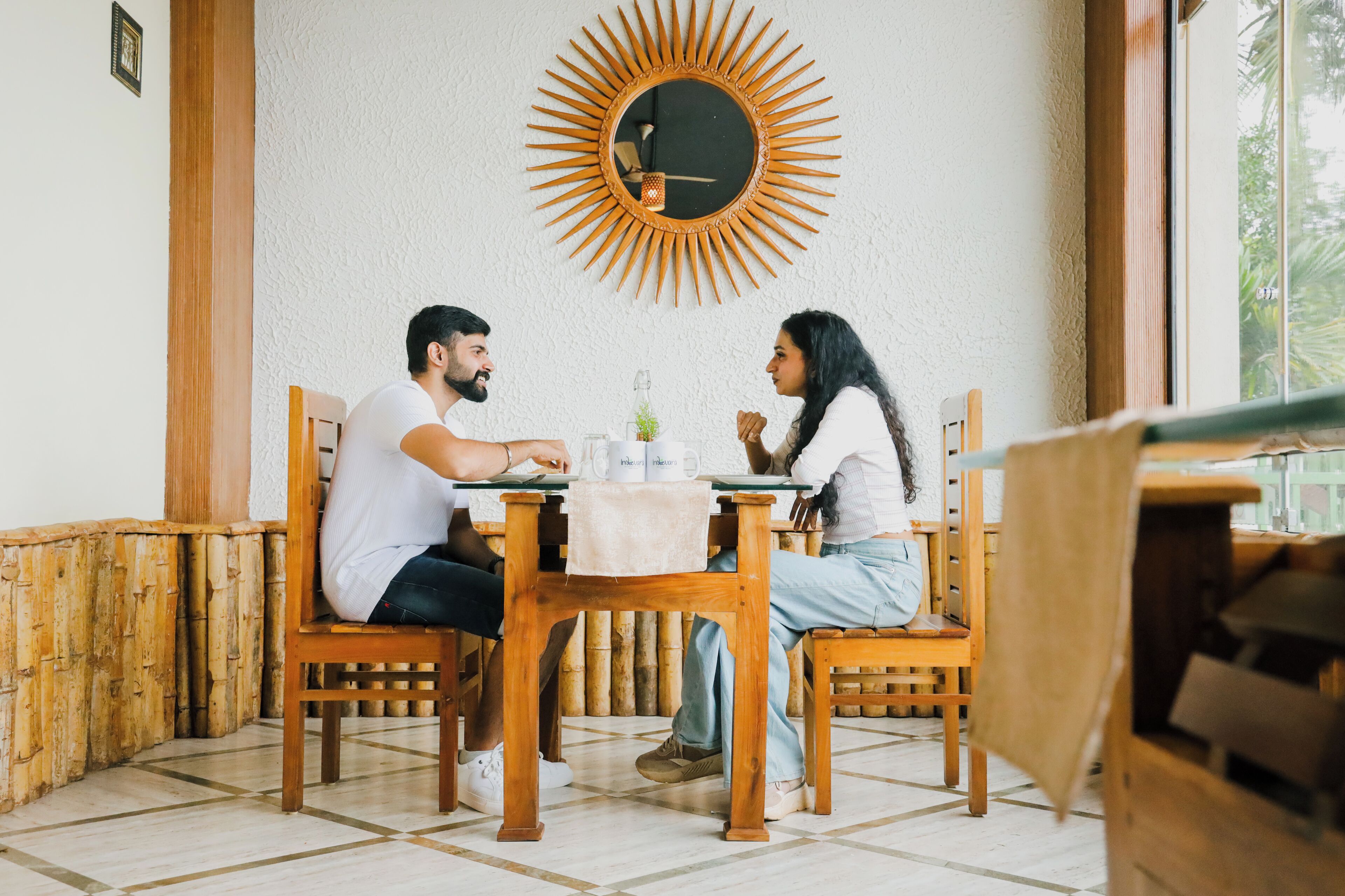 A dining area with a wooden table and chairs, a round mirror with a sunburst design, and a window with a view of greenery.