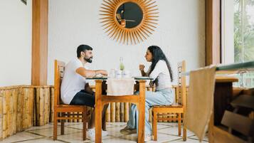 A dining area with a wooden table and chairs, a round mirror with a sunburst design, and a window with a view of greenery.