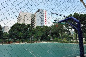 Ein Basketballplatz im Freien mit einem Maschendrahtzaun, Bäumen und einem Gebäude im Hintergrund.