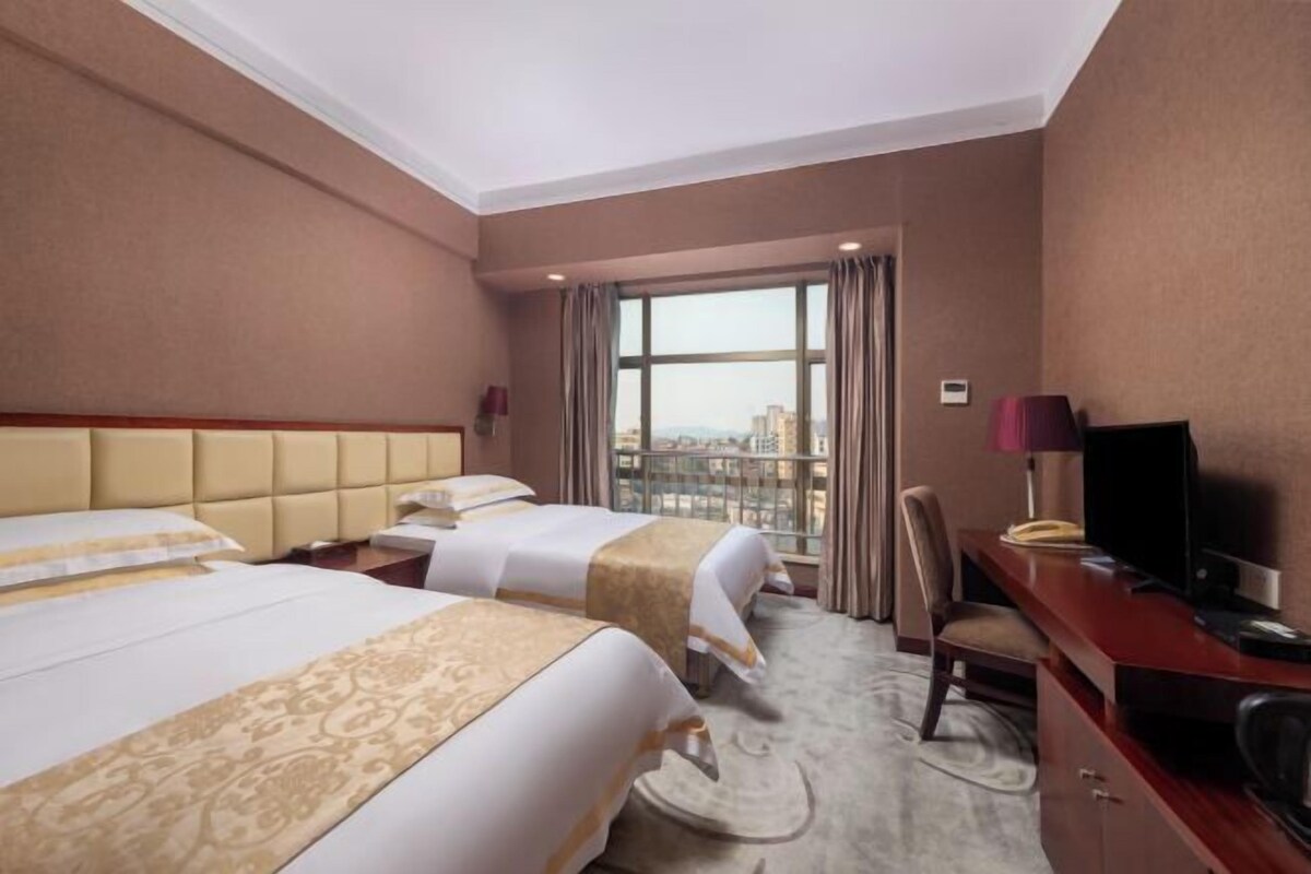A hotel room with two beds, a desk, a television, and a view of the city through large windows.