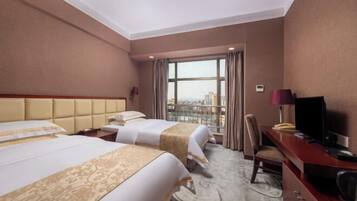 A hotel room with two beds, a desk, a television, and a view of the city through large windows.