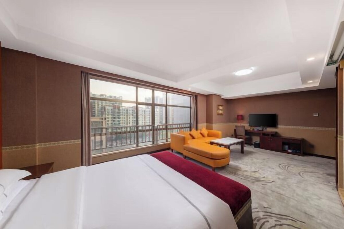 A hotel room with a large bed, a sofa, a TV, and a view of the city through a large window.