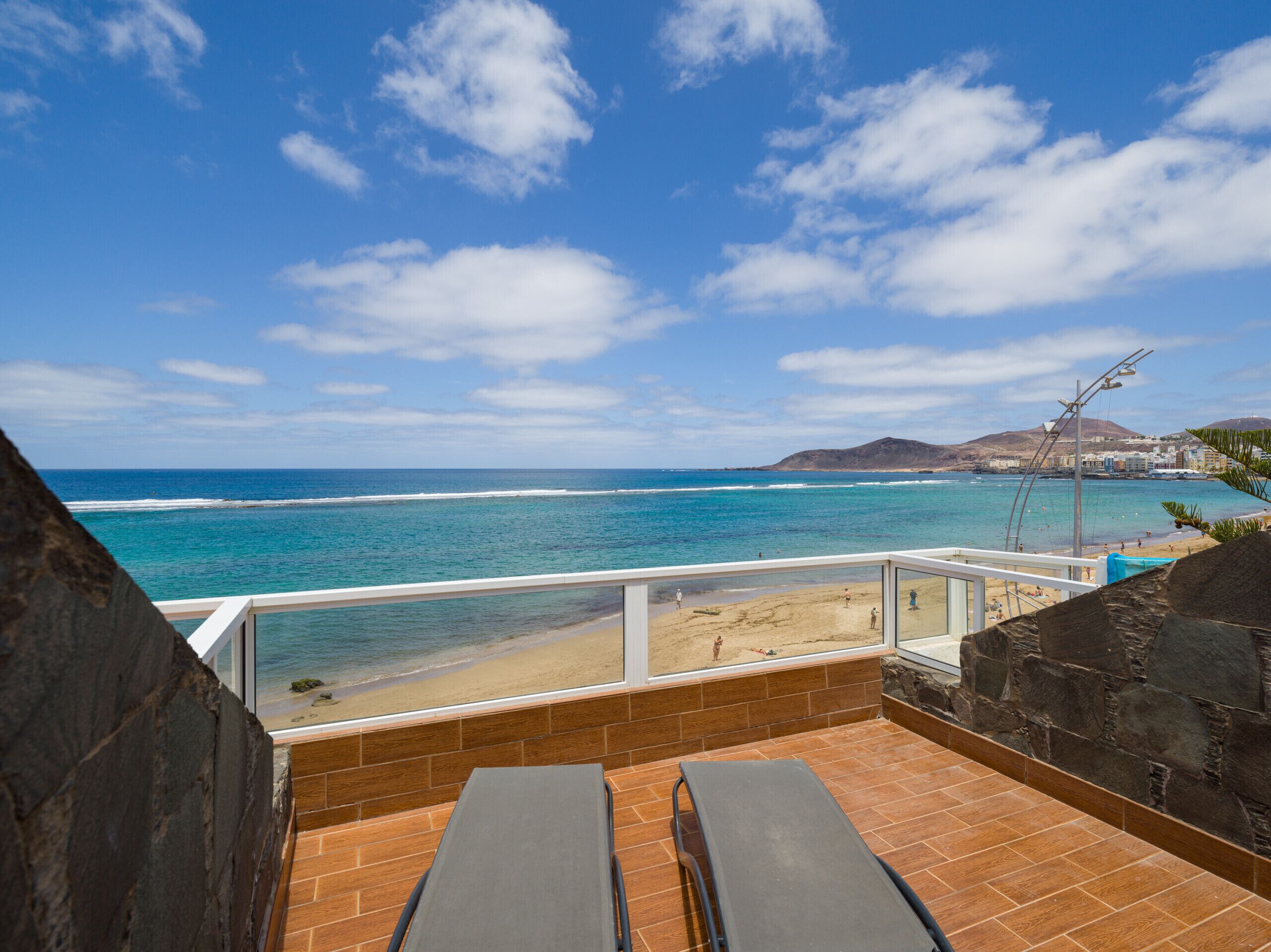 A balcony with a view of the ocean and beach, featuring two lounge chairs and a stone wall.