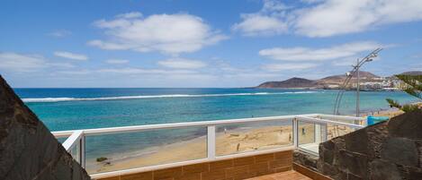 A balcony with a view of the ocean and beach, featuring two lounge chairs and a stone wall.