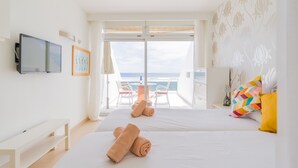 A hotel room with two beds, a TV, a desk, and a balcony with a view of the sea.