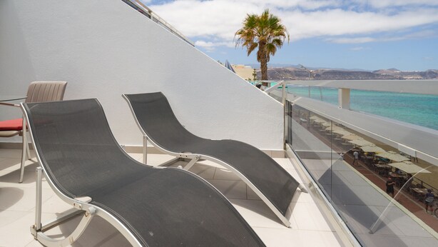 A balcony with two lounge chairs, a palm tree, and a view of the sea.