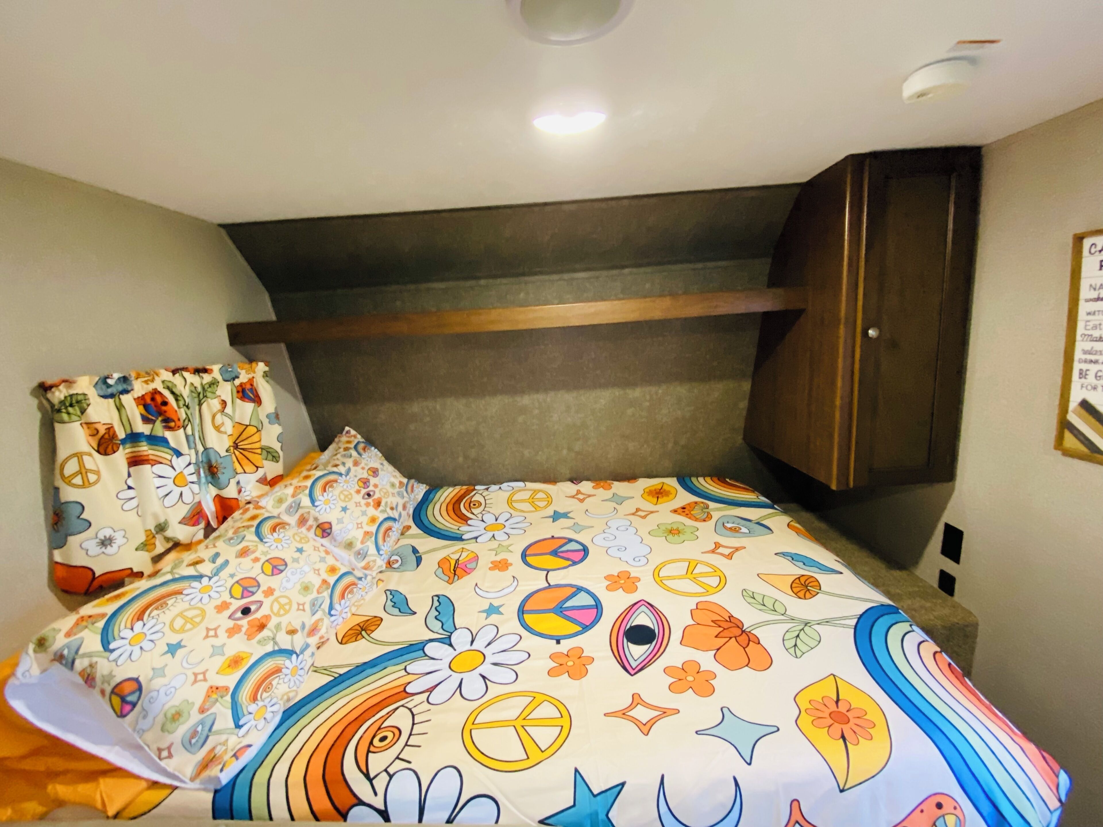 A bed with a colorful floral and peace symbol bedspread, a wooden headboard, and a dark wooden wardrobe.