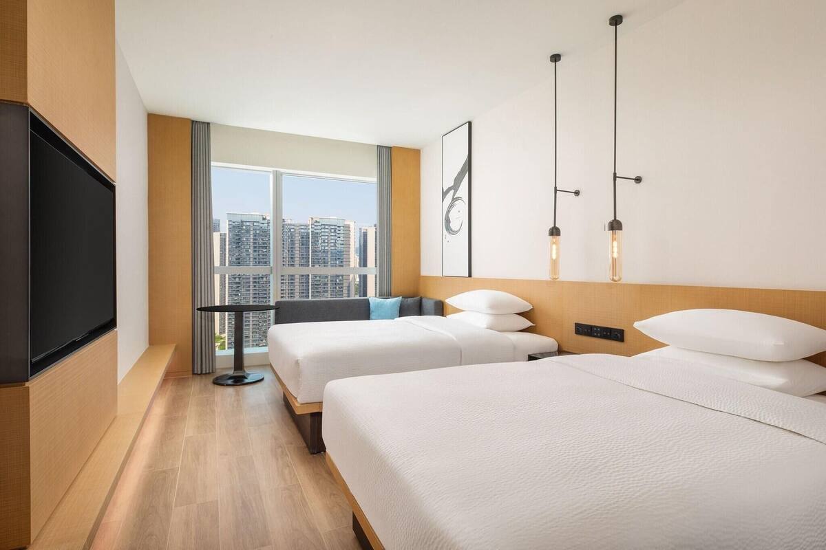 A hotel room with two beds, a TV, a table, and a large window offering a city view.