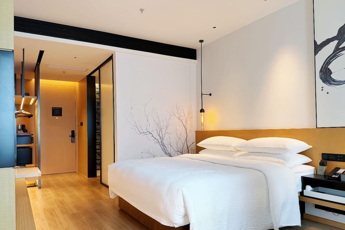 A modern hotel room with a large bed, a wooden floor, a wardrobe, and a desk with a chair.