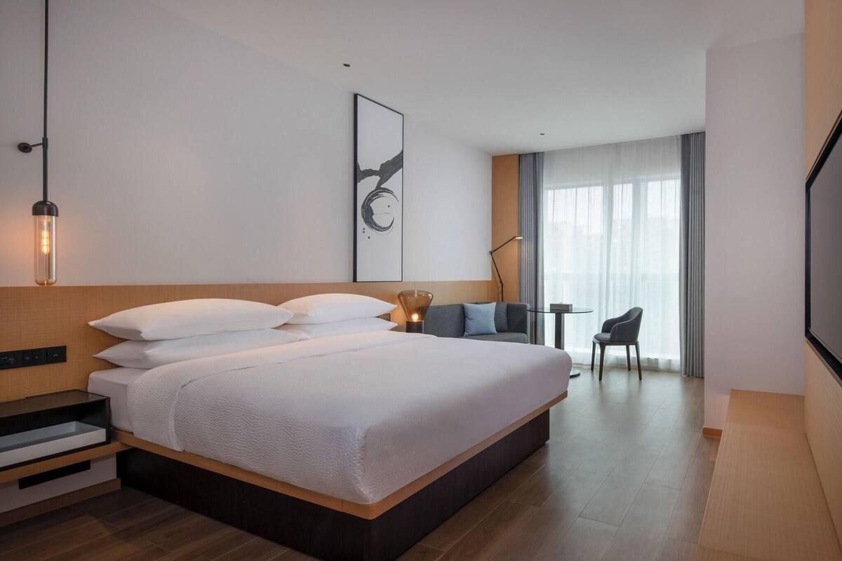 A modern hotel room with a large bed, a desk, a chair, and a large window with curtains.