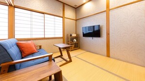Una habitación japonesa tradicional con suelo de tatami, una mesa de madera baja, un sofá con cojines, un televisor y una estantería con objetos.