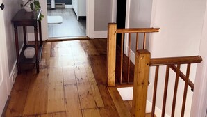 Una escalera de madera con barandilla que lleva a un rellano en el segundo piso. Se ve una pequeña mesa con una planta en maceta a la izquierda.