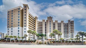 A multi-story hotel with a central courtyard, palm trees, and a clear sky. - Daytona Beach Condo: Pool Access, Oceanfront (Daytona Beach Shores)