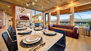 Un espace repas avec une longue table dressée pour un repas, entourée de chaises, et offrant une vue sur les montagnes grâce à de grandes fenêtres.