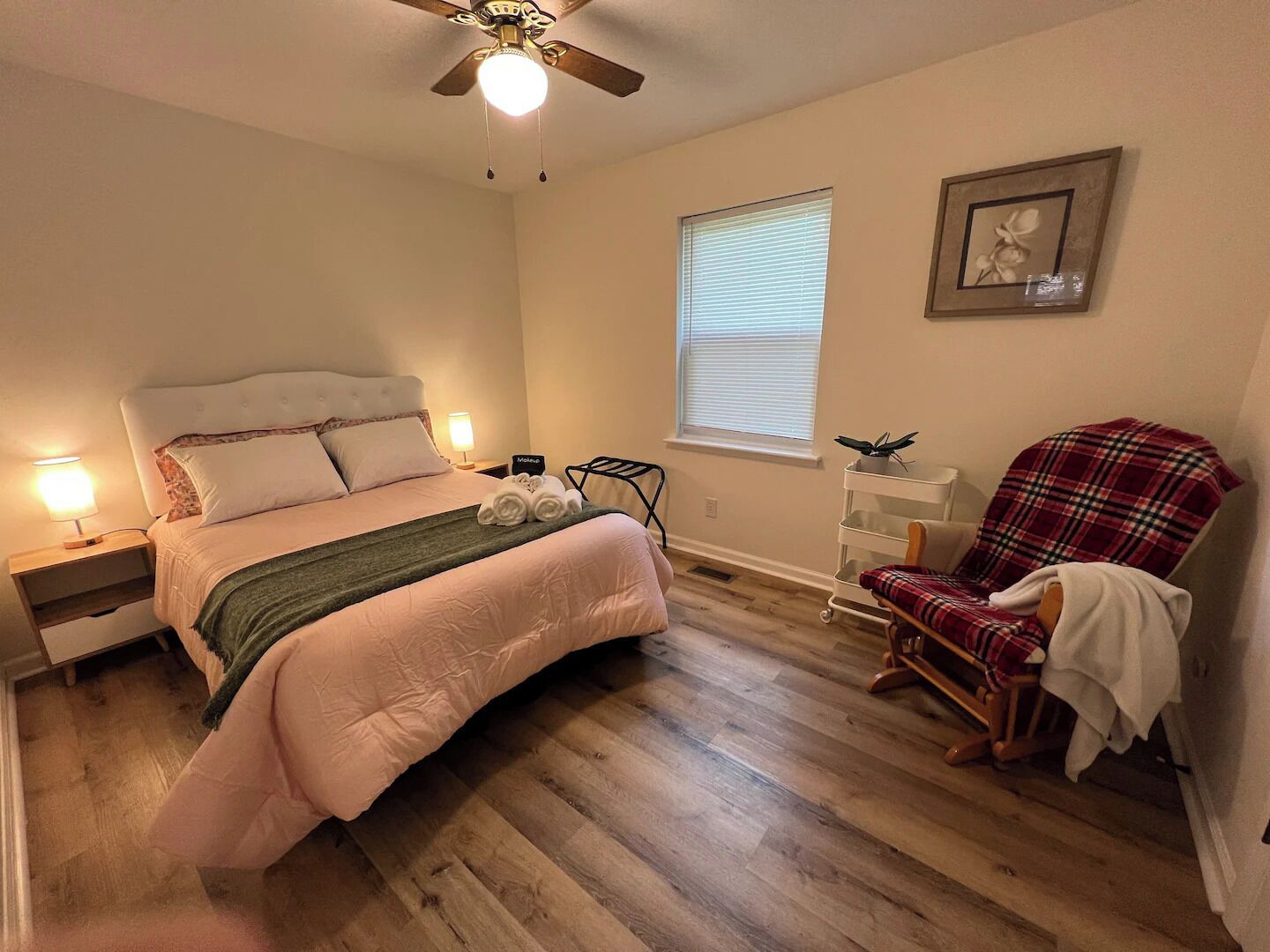 A bedroom with a bed, a chair, a nightstand, a bedside table, a ceiling fan, and a window with blinds.