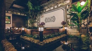 Ein traditioneller chinesischer Garten mit einer Holzbrücke, einem Steinbrunnen und üppiger Vegetation.