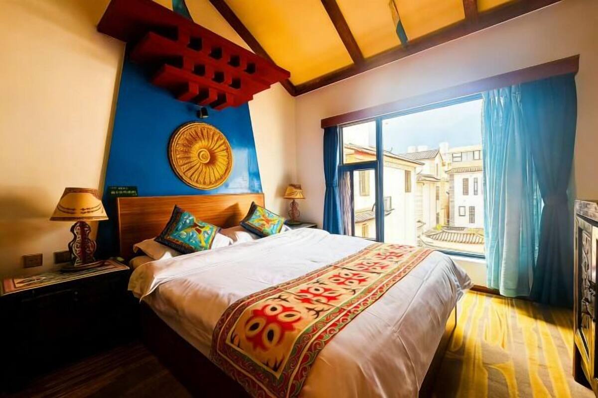 A hotel room with a bed, nightstands, a decorative wall, and a view of buildings through the window.