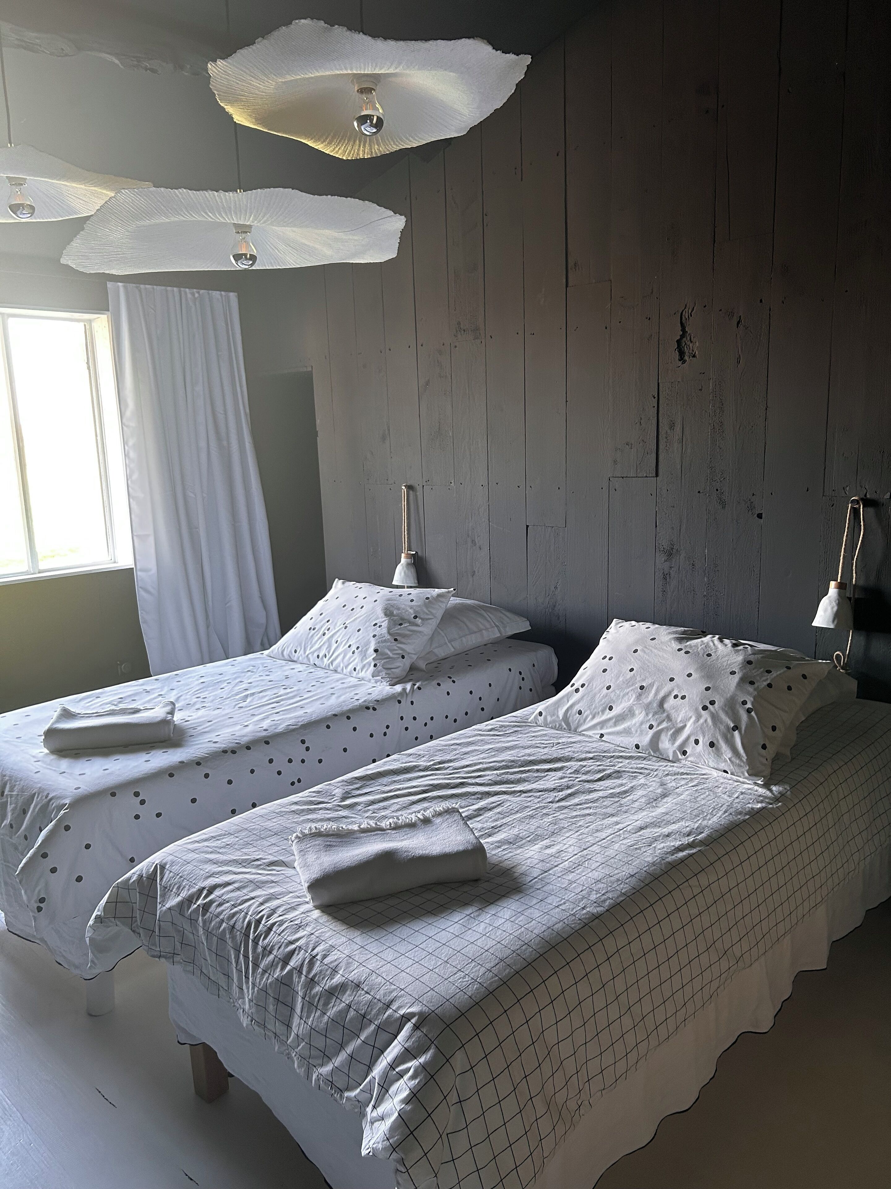 Une chambre avec deux lits simples, chacun équipé d’un oreiller et d’une serviette pliée. On y trouve une fenêtre avec des rideaux blancs, un mur en bois et un ventilateur au plafond.