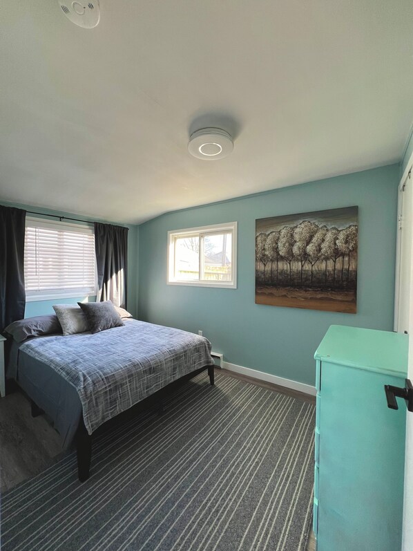 A bedroom with a bed, a window, a painting, and a teal cabinet.