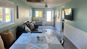 A modern living room with a sofa, a dining table with wine glasses, a ceiling fan, and a TV mounted on the wall. - Little Blue on Second St! Cozy & close to the beach, pub, bar, and downtown! (Port Stanley)