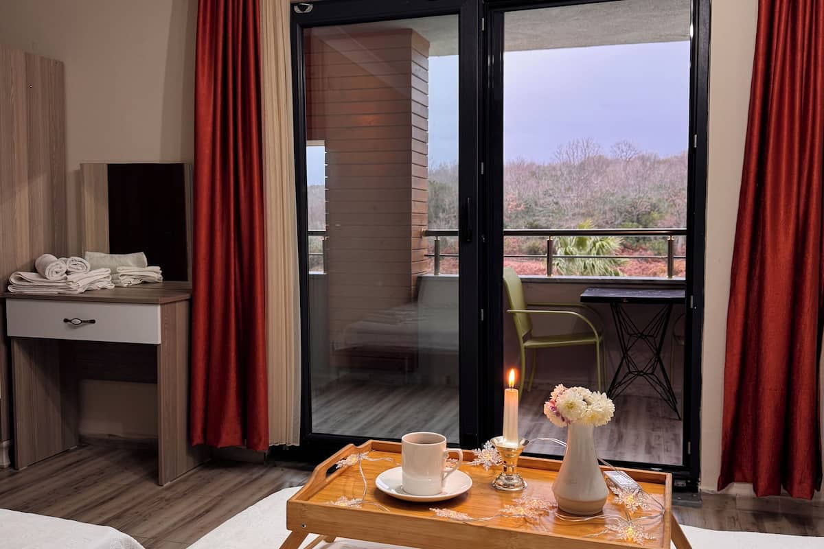 Une chambre d’hôtel avec un lit, un bureau, une table avec une bougie, un vase de fleurs et une vue sur l’extérieur.