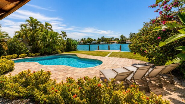 A swimming pool with a curved edge, surrounded by a patio area with lounge chairs, and a view of a body of water with buildings in the background.