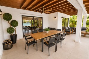 Een overdekt buitendinergedeelte met een houten tafel en stoelen, planten in potten en uitzicht op een zwembad.