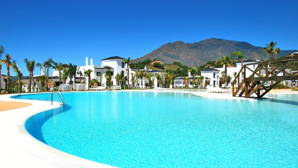 A large, clear blue swimming pool with palm trees and white buildings in the background.