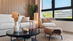 Un salon moderne comprenant un canapé, une table basse ronde, un fauteuil en osier et une grande fenêtre donnant sur des bâtiments et de la verdure.