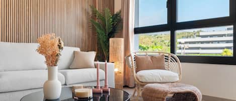 A modern living room with a sofa, a round coffee table, a wicker chair, and a large window offering a view of buildings and greenery.