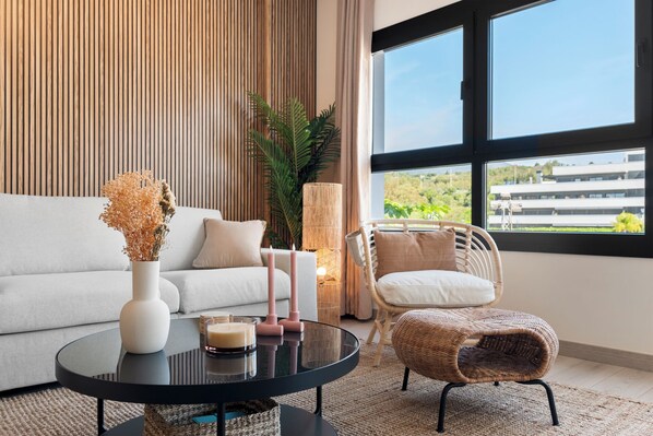 A modern living room with a sofa, a round coffee table, a wicker chair, and a large window offering a view of buildings and greenery.