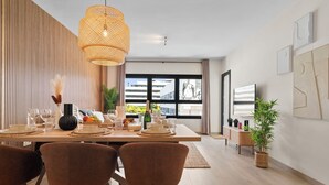 Un espace repas moderne avec une table en bois mise pour le petit-déjeuner, agrémenté d’un luminaire suspendu en fibres tressées, de chaises rembourrées et offrant une vue sur la ville grâce à de grandes fenêtres.