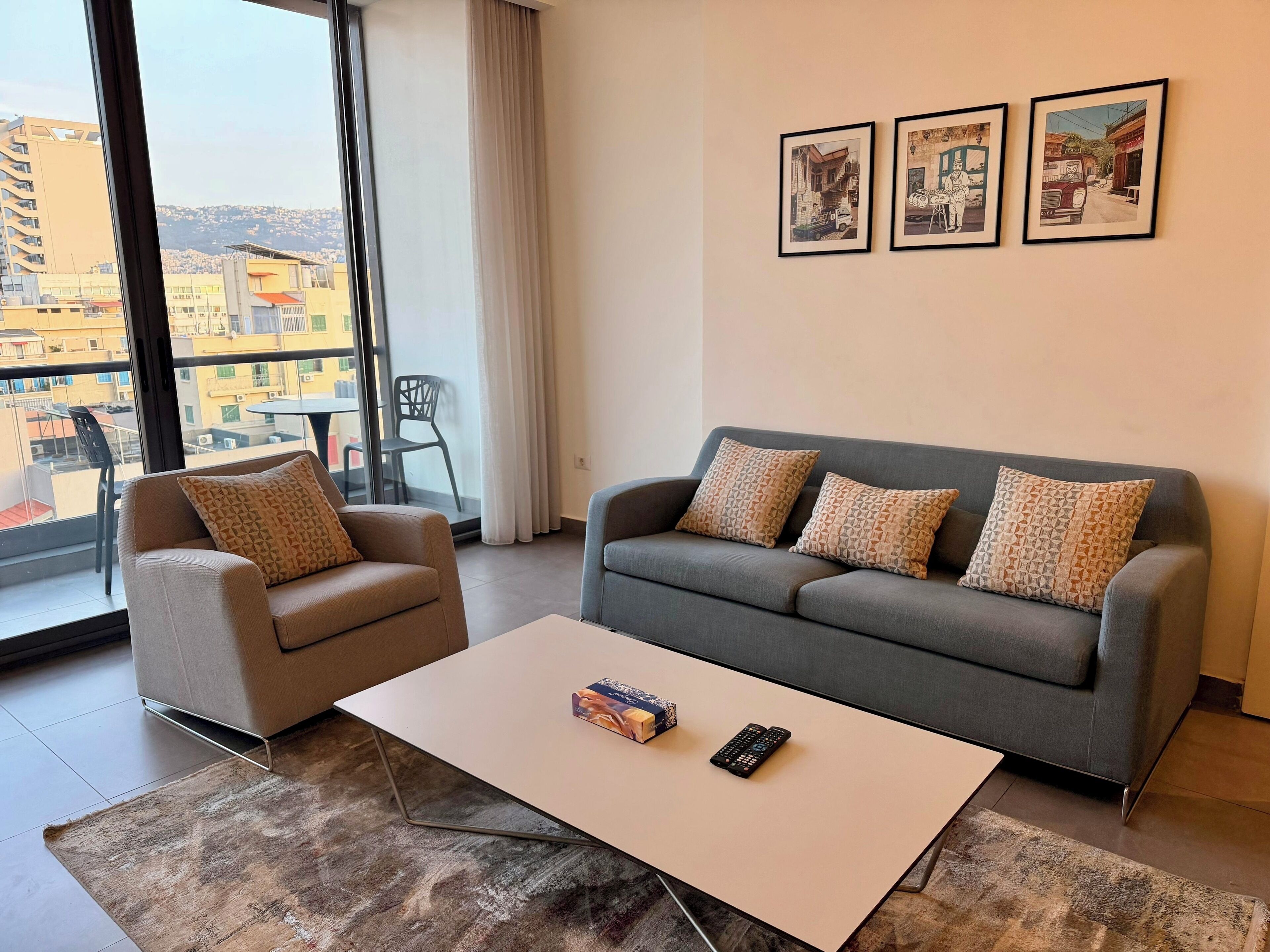 A modern living room with a grey sofa, beige armchairs, and a white coffee table. There are three framed pictures on the wall, and a balcony with a view of buildings is visible through the window.