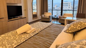 A hotel room with a large bed, two armchairs, a small table, a TV, and a view of the city through large windows. - Unit for Living (Beirut)